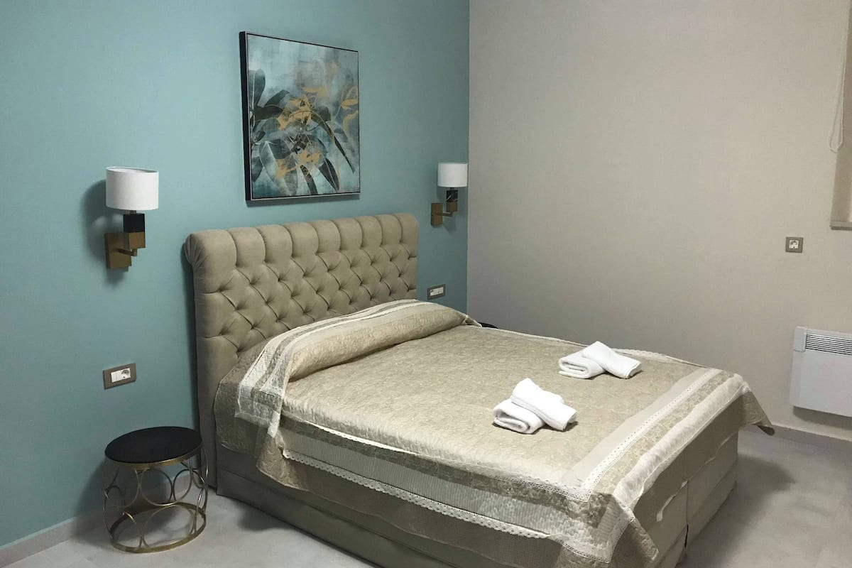 A neatly made bed with a tufted headboard, two bedside lamps, a painting on the wall, and a radiator.