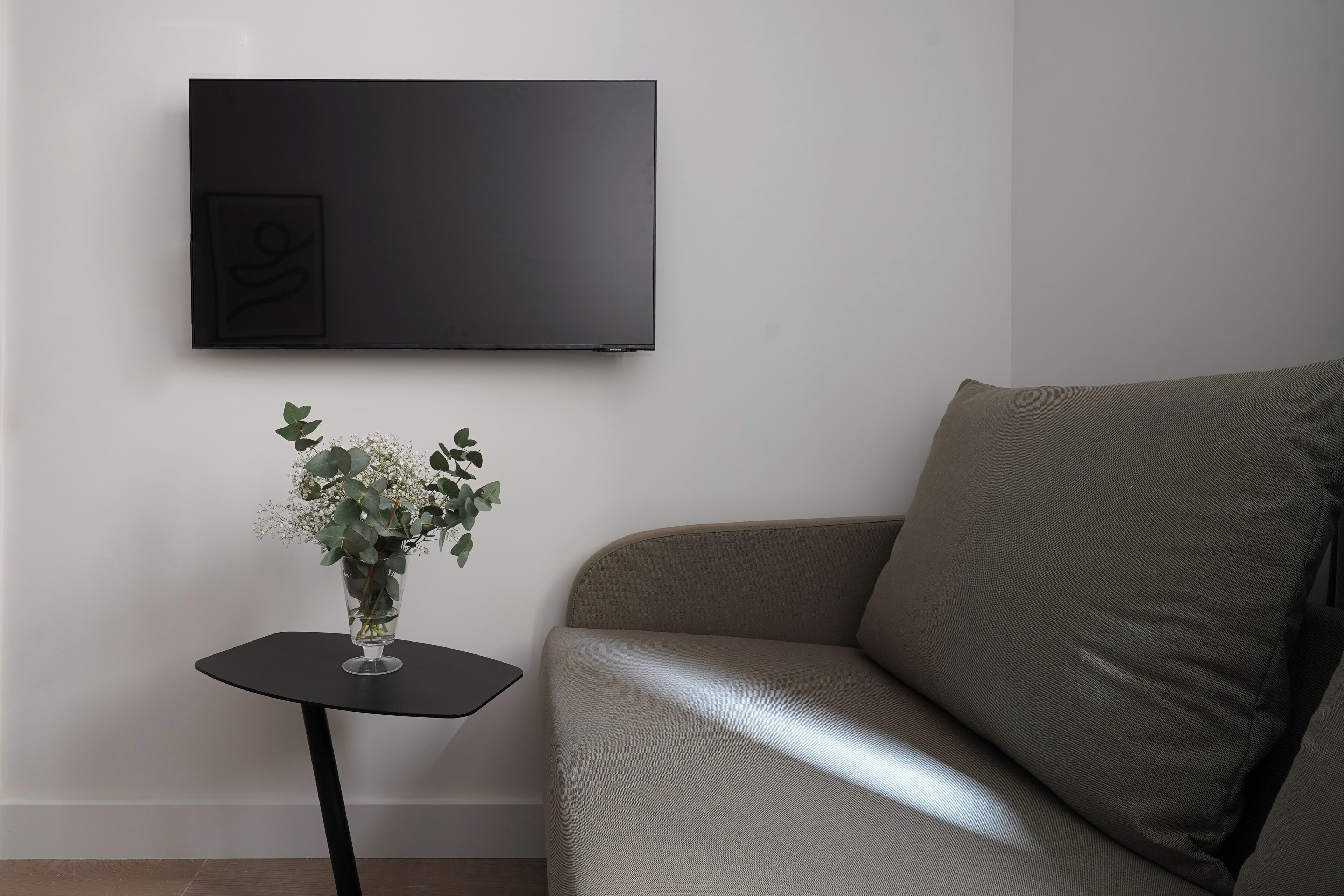 A living room with a flat-screen TV mounted on the wall, a small black side table with a vase of flowers, and a beige sofa.