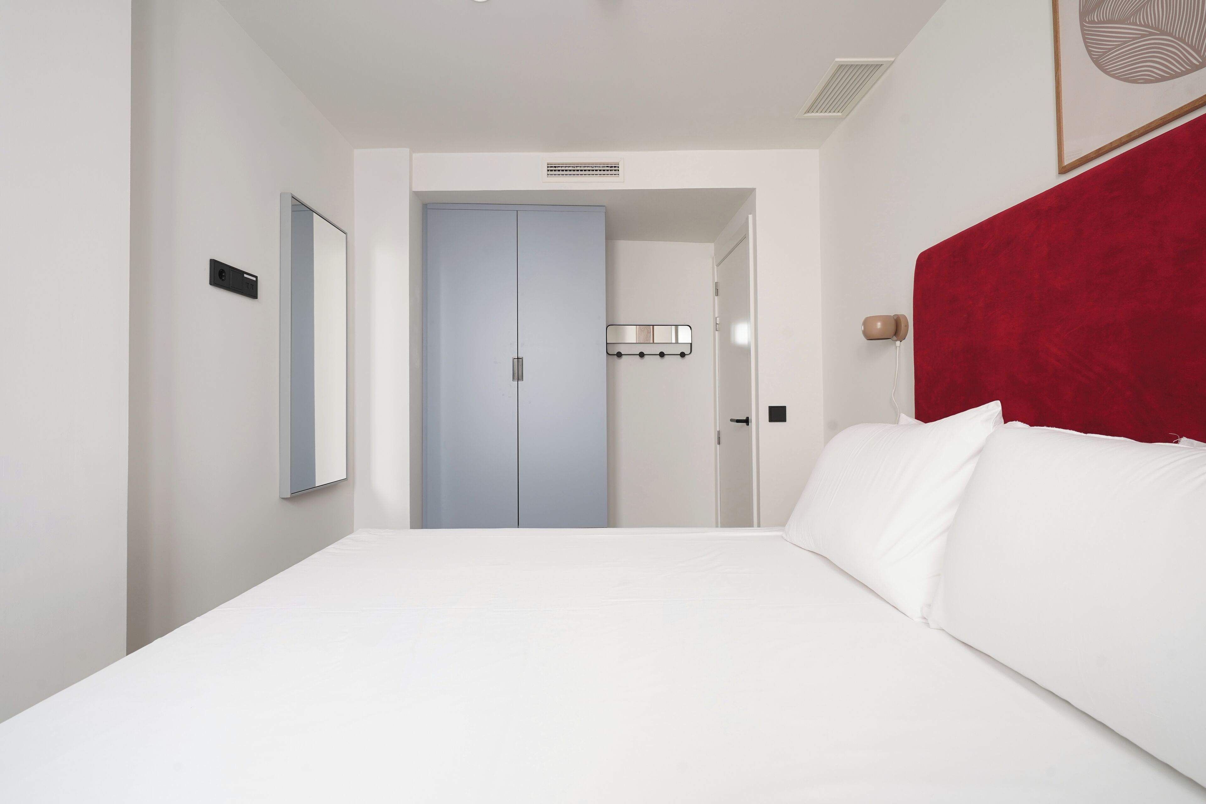 Habitación de hotel con una cama grande, cabecera roja, puerta azul claro y una lámpara fijada a la pared.