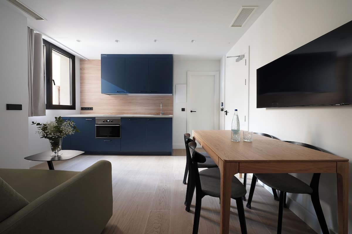 Una cocina moderna con armarios azules, encimera de madera y horno empotrado. Hay una mesa de comedor con sillas, un florero con flores y un televisor de pantalla plana montado en la pared.