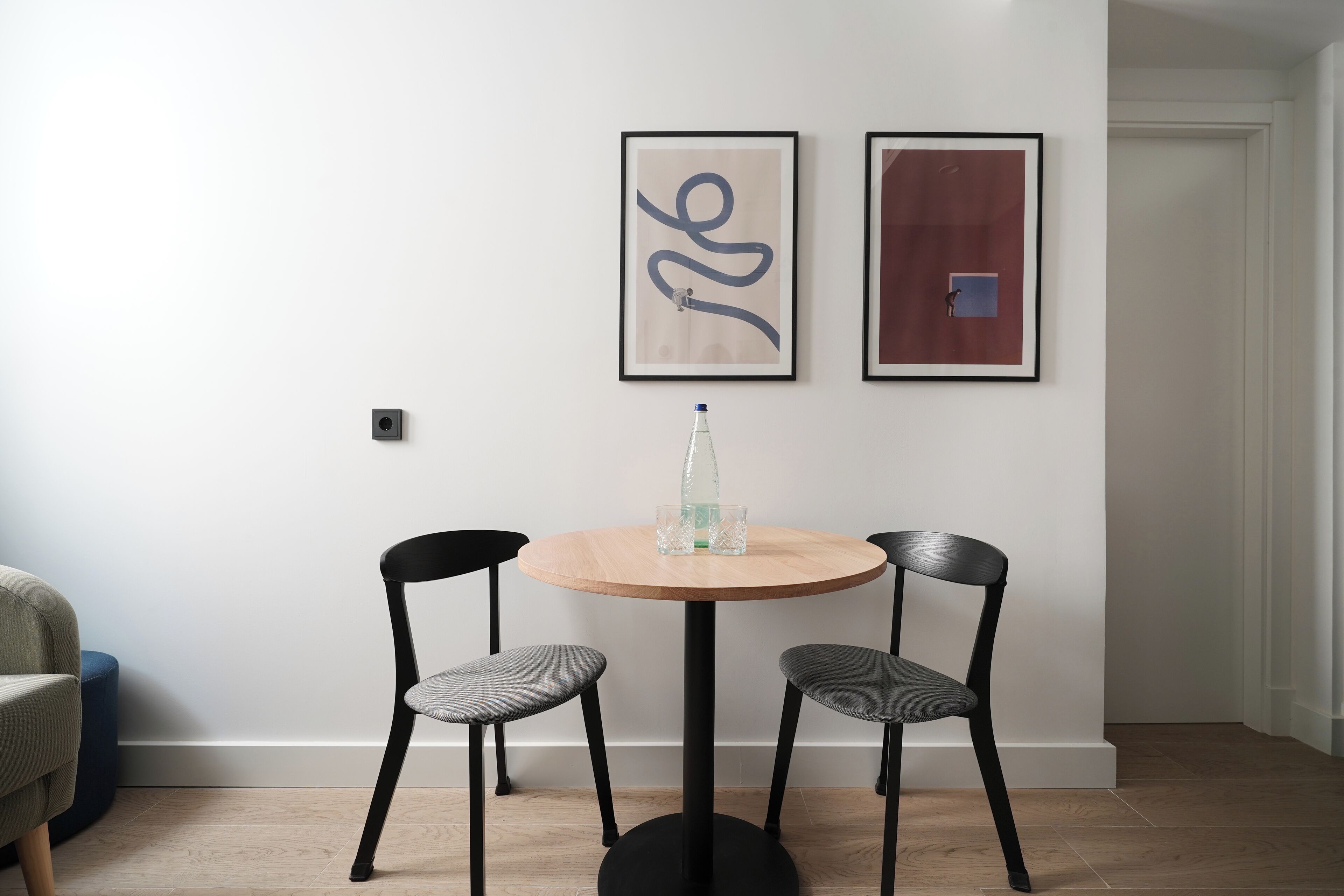 Un comedor con una mesa redonda de madera, dos sillas negras y dos obras de arte enmarcadas en la pared.