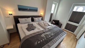 A bedroom with a bed, pillows, a blanket, a bedside table, a chair, a window, and a painting on the wall. - Meerzeit - im Kaiserhof Wangerooge (Wangerooge)