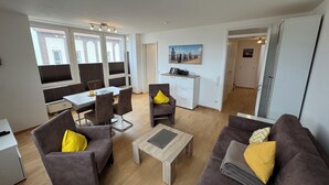 A modern living room with a dining area, featuring a sofa set, a coffee table, and a dining table with chairs. There is a white cabinet with drawers and a painting on the wall. - Meerzeit - im Kaiserhof Wangerooge (Wangerooge)