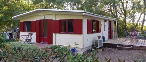 A small, single-story building with red shutters, a door, and windows. It has a wooden deck with a table and chairs.