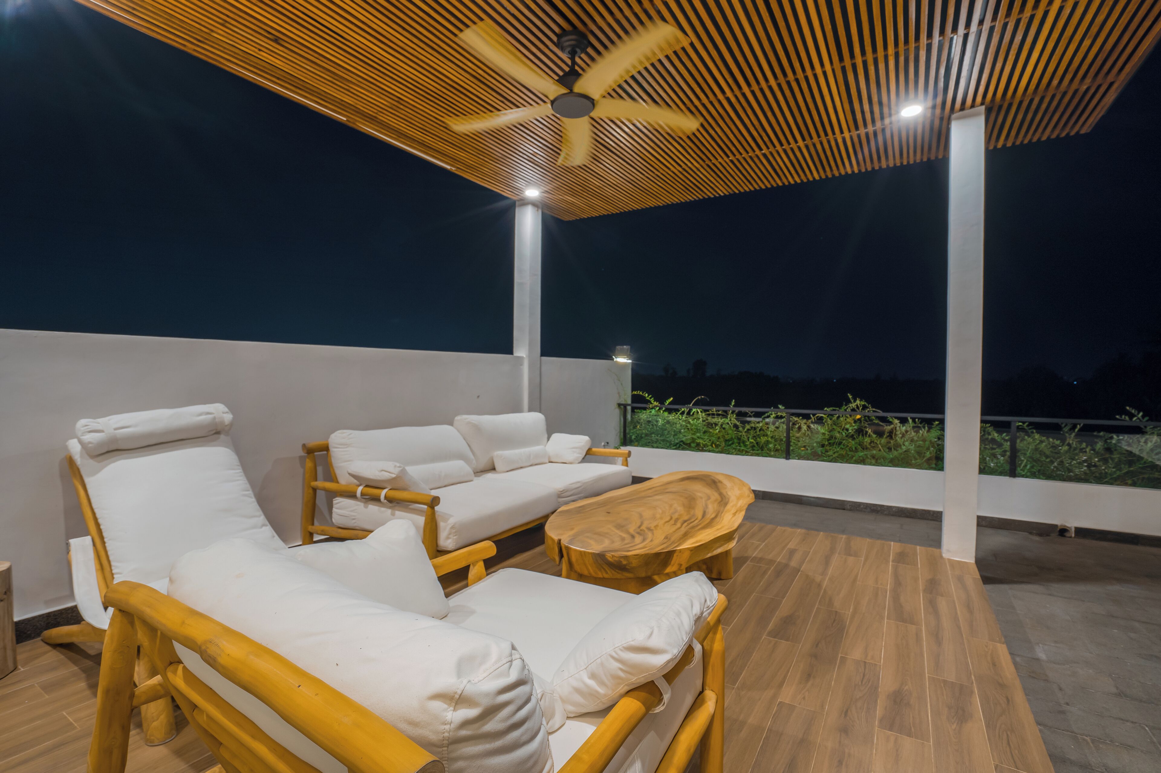 Een terras op het dak met houten vloer, witte loungestoelen met kussens, een houten salontafel en een plafondventilator.