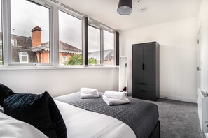 A bedroom with a bed, two windows, a wardrobe, and a view of a building outside. - Chic 1BR Wrexham Apt, Walk to Station (Wrexham)