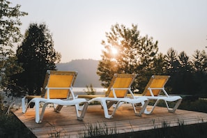 Two white lounge chairs with yellow cushions on a wooden deck overlooking a body of water with trees in the background.