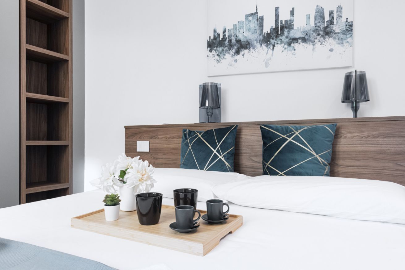Un lit bien fait, avec une tête de lit en bois, une œuvre d’art représentant un paysage urbain accrochée au mur, et un plateau avec des tasses et une plante posé sur la table de chevet.