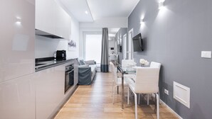 Et moderne køkken med hvide skabe, en sort bordplade og et spiseområde med et glasbord og hvide stole.