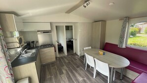 A compact kitchen with a gas stove, sink, and microwave. There is a dining area with a table and chairs, and a red sofa. The room has a window with curtains. - XL Mobile-home for 6 -Pommeraie de l'Océan- 4Star (Trégunc)