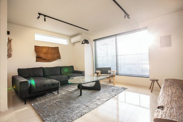 A modern living room with a glass coffee table, a dark sofa, and a large window with blinds.