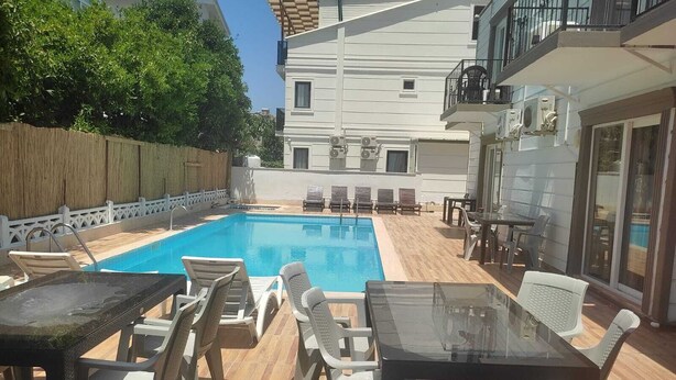 A pool area with lounge chairs and tables, a wooden deck, and a building with balconies in the background. - GARDEN Prizma Pansiyon & Aparts (Kemer)