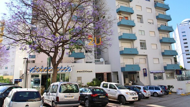 A multi-story building with balconies, a parking area with several cars, and a tree with purple flowers.
