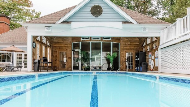 A swimming pool with a wooden deck area, a covered seating area, and a building with a clock and a sign.