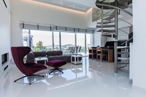 Una sala de estar moderna con un ventanal grande, una escalera curva y una zona de estar con un sillón y un reposapiés rojos.