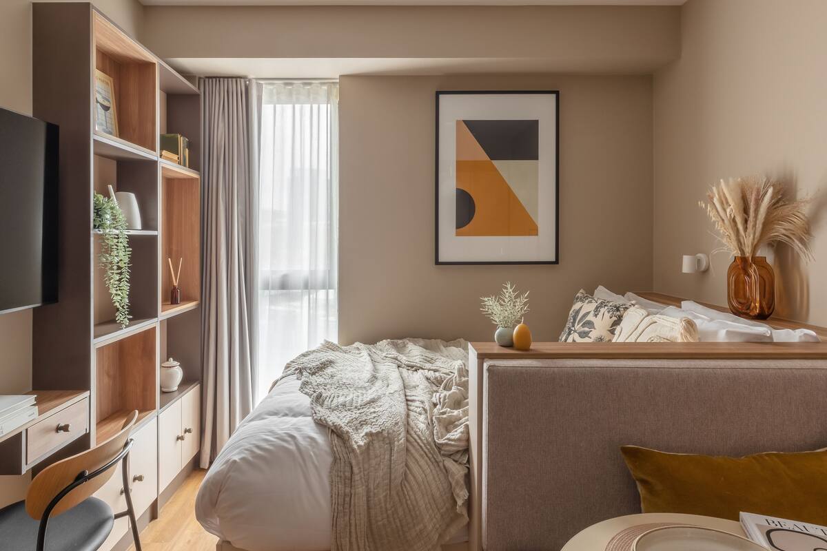 A modern hotel room with a bed, a desk, a bookshelf, and a large window with curtains.