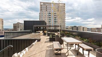 Una terraza en la azotea con cubierta de madera, zonas de estar y vista a un edificio alto al fondo.