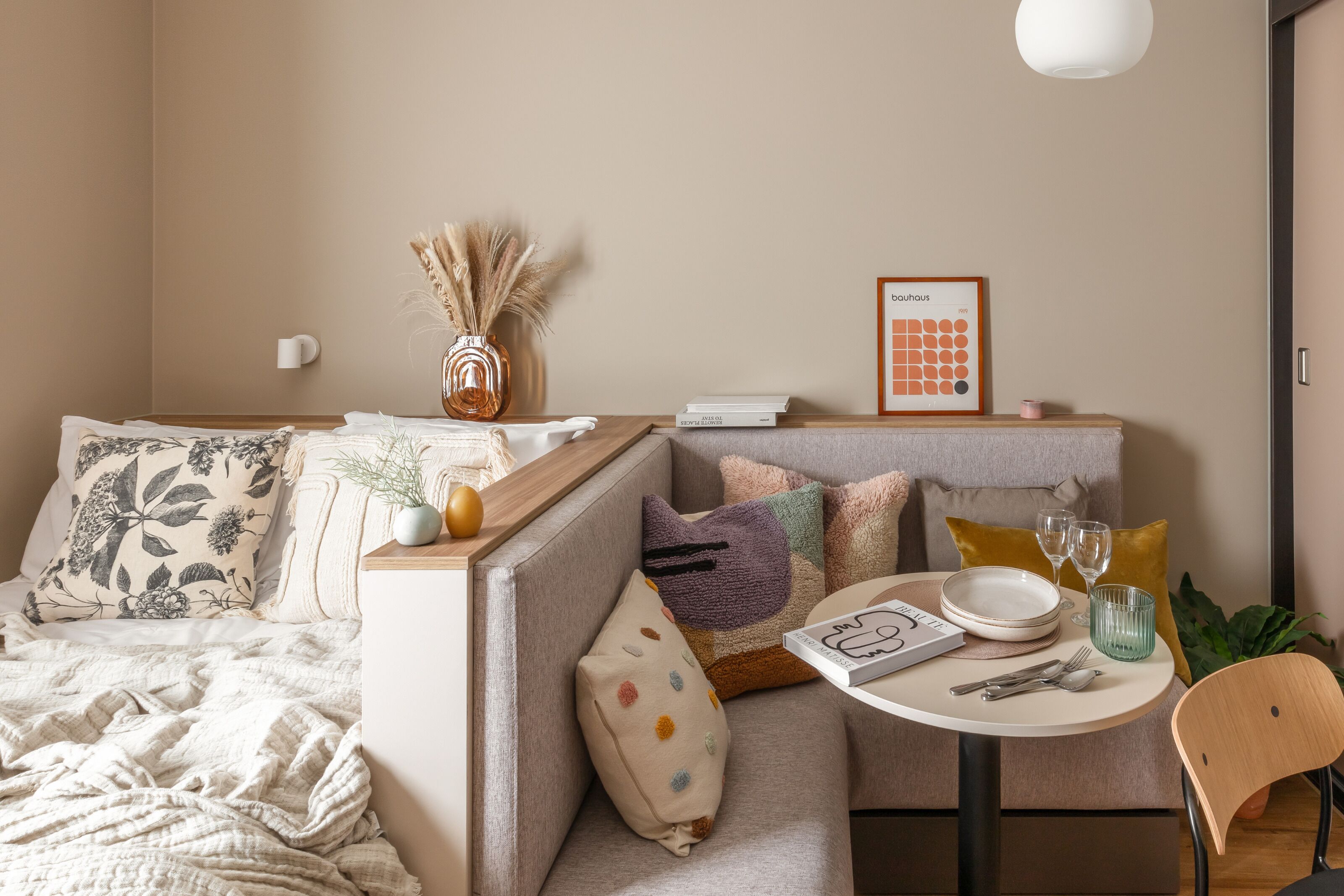 A cozy room with a sofa, a small dining table with plates and glasses, a vase with dried plants, and a framed picture on the wall.