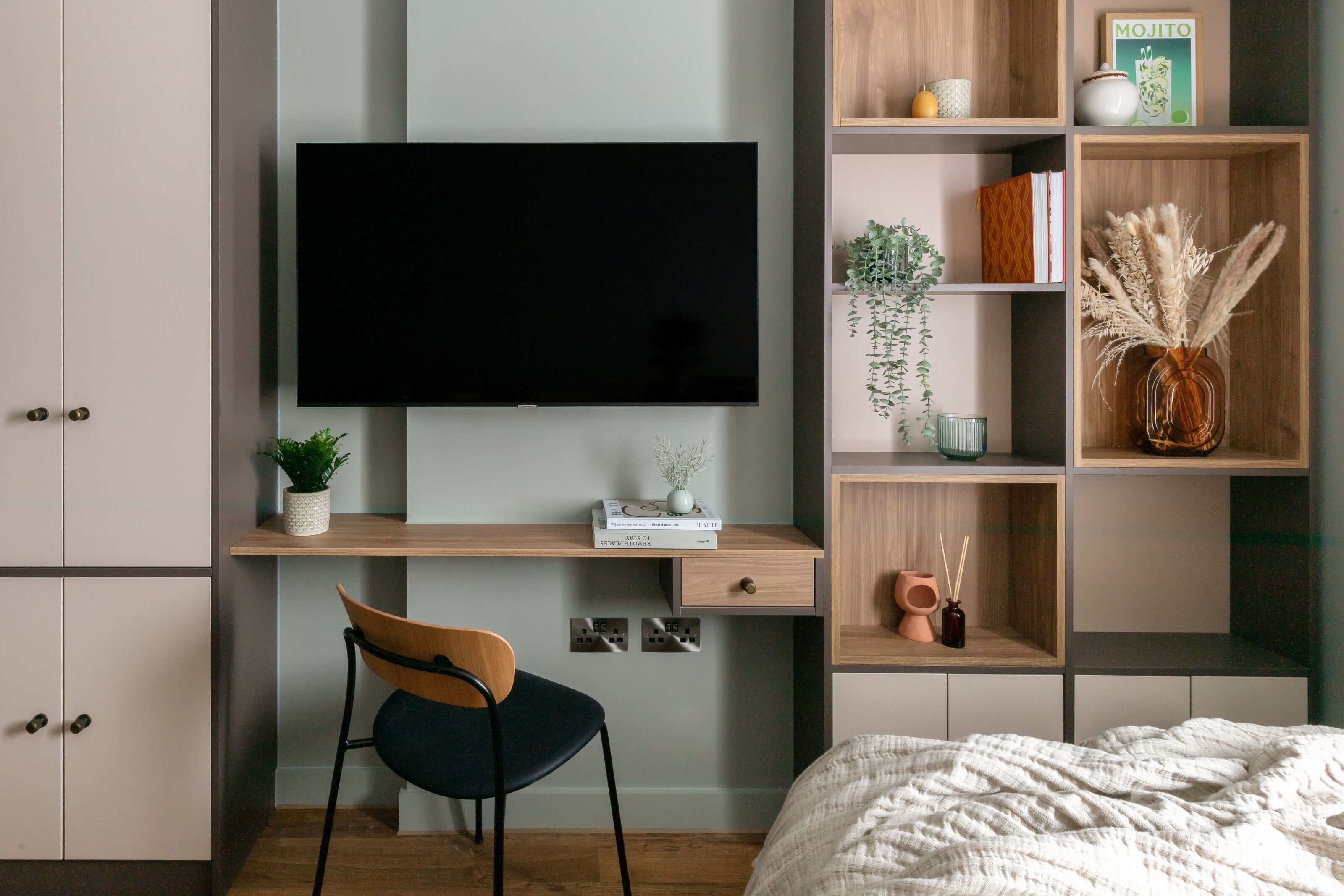 A bedroom with a large flat-screen TV mounted on the wall, a wooden desk with a chair, a bed with a white comforter, and a bookshelf with various items.