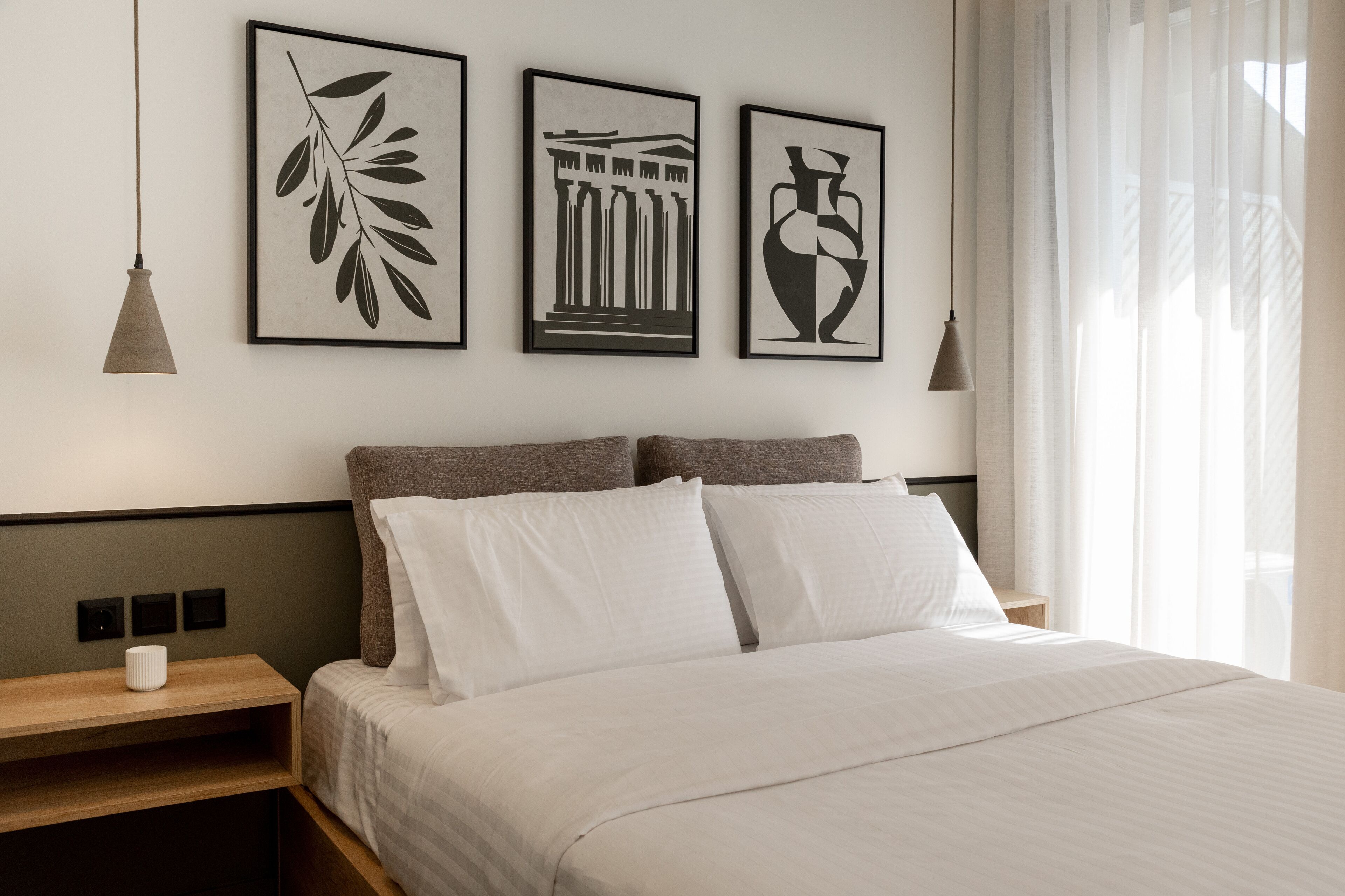 Una cama bien hecha con sábanas blancas, una mesita de noche de madera con un florero blanco, y tres cuadros en la pared.