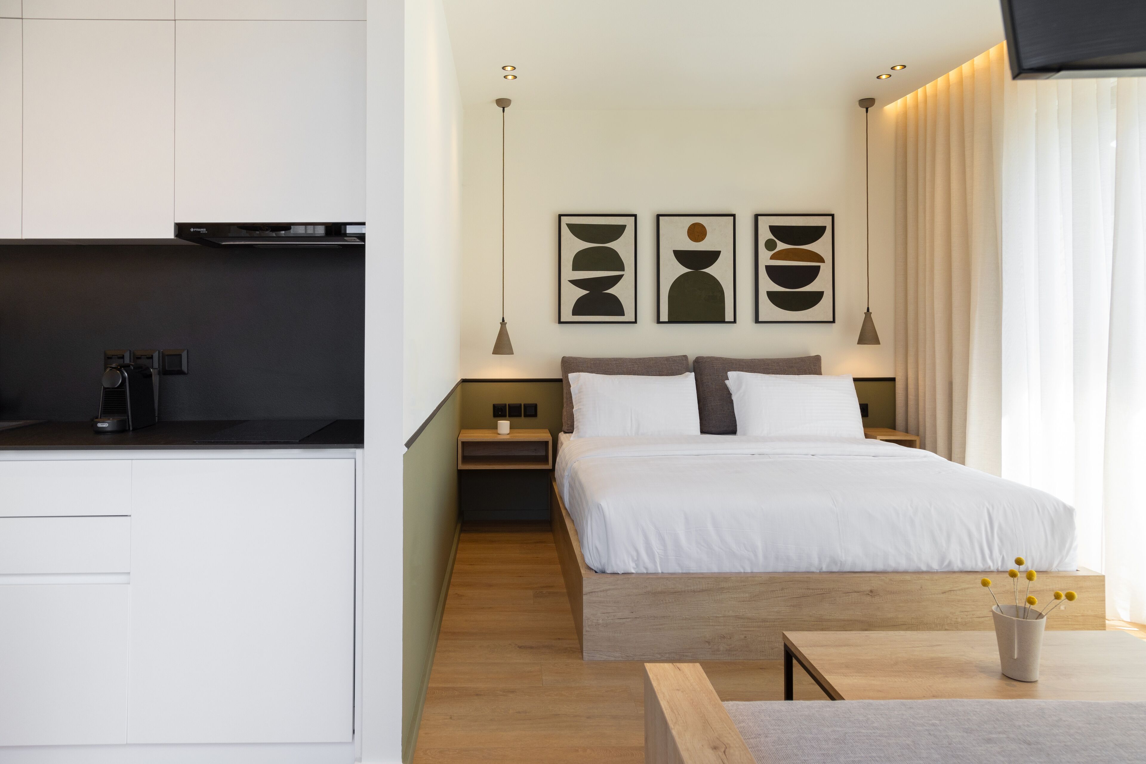 A modern hotel room with a bed, bedside tables, a small desk, and a kitchenette.