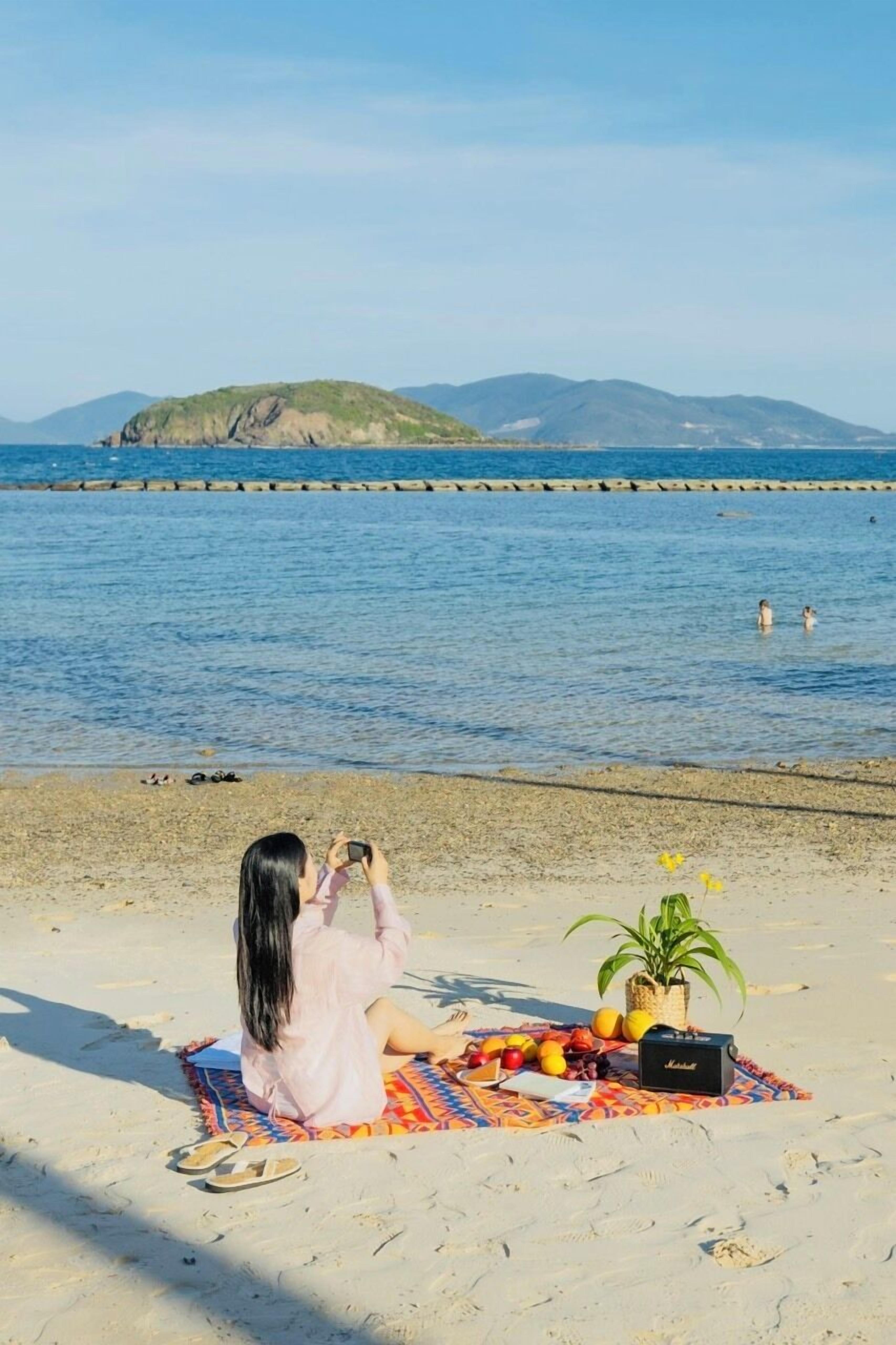 A beach scene with a person sitting on a blanket, taking a photo. There is a picnic setup with fruits, a plant, and a speaker on the blanket.