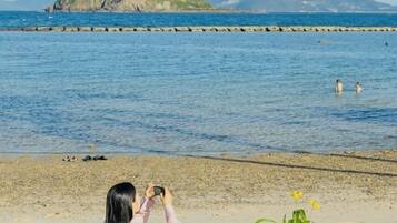 A beach scene with a person sitting on a blanket, taking a photo. There is a picnic setup with fruits, a plant, and a speaker on the blanket.
