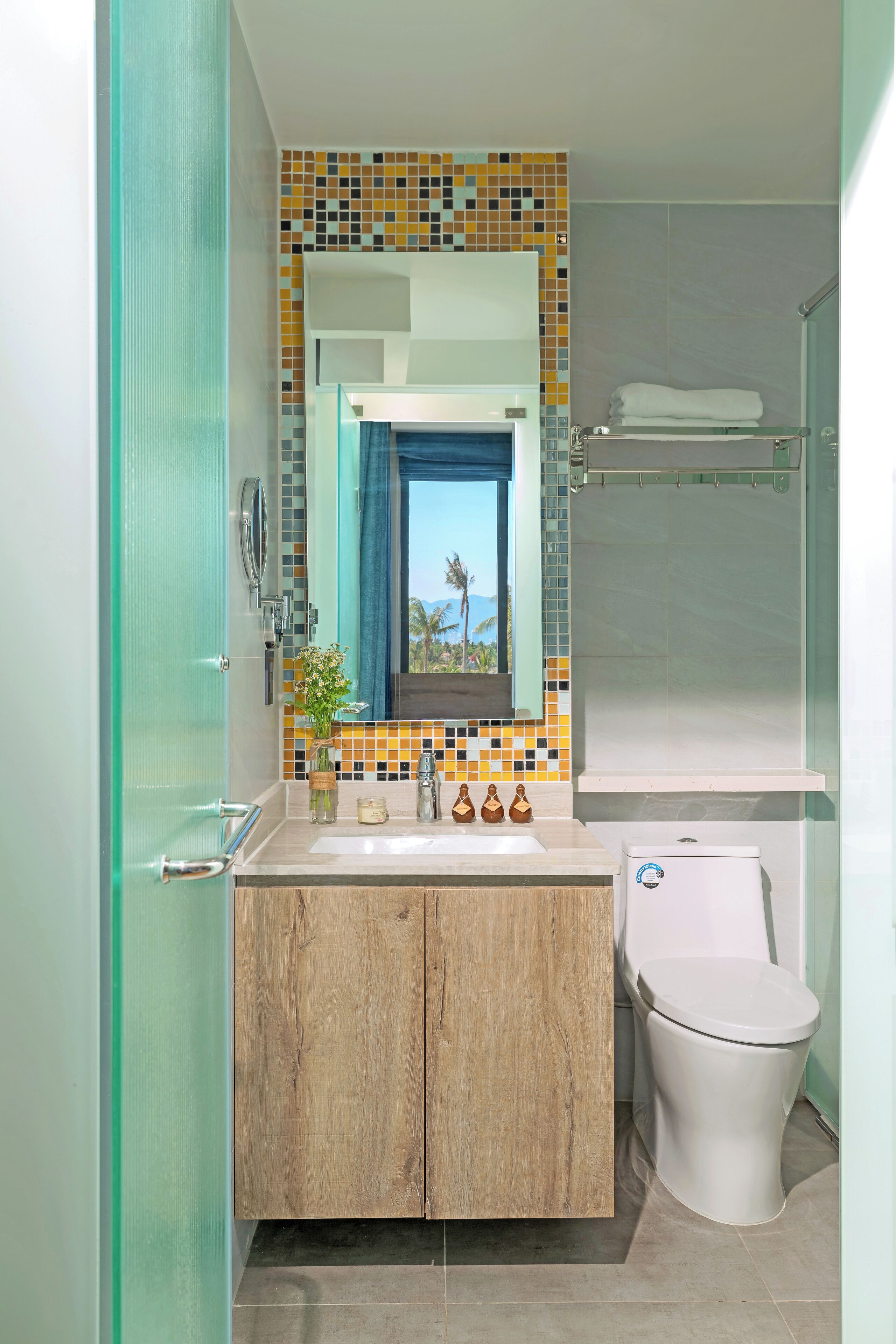 Een badkamer met een houten wastafelmeubel, een wit toilet, een betegelde spatwand en een raam met uitzicht op palmbomen.