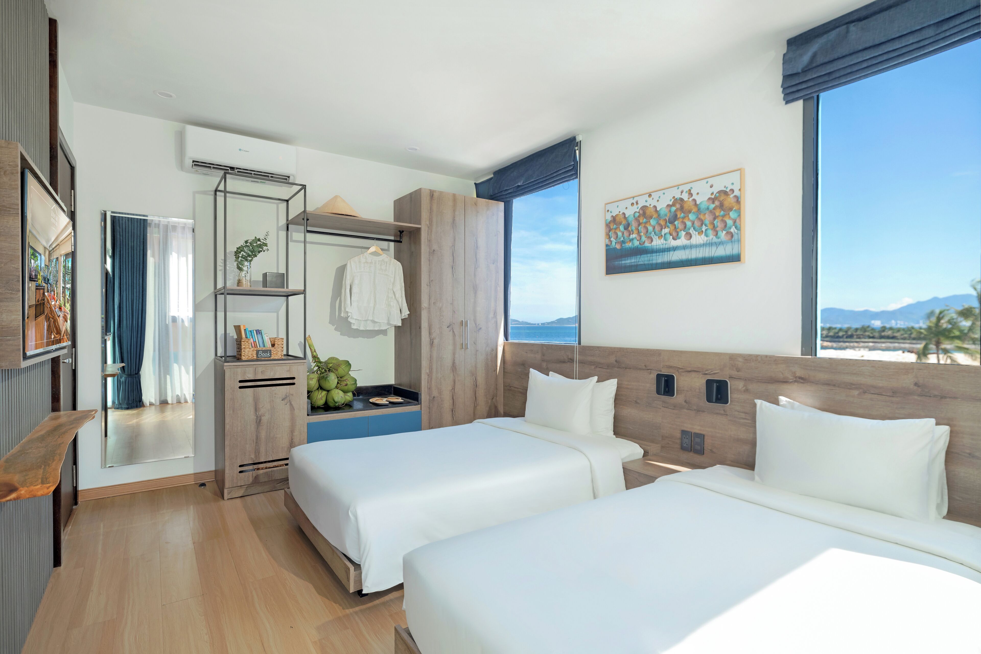 A modern hotel room with two beds, a wardrobe, a vanity with a mirror, and a large window offering a view of the mountains.
