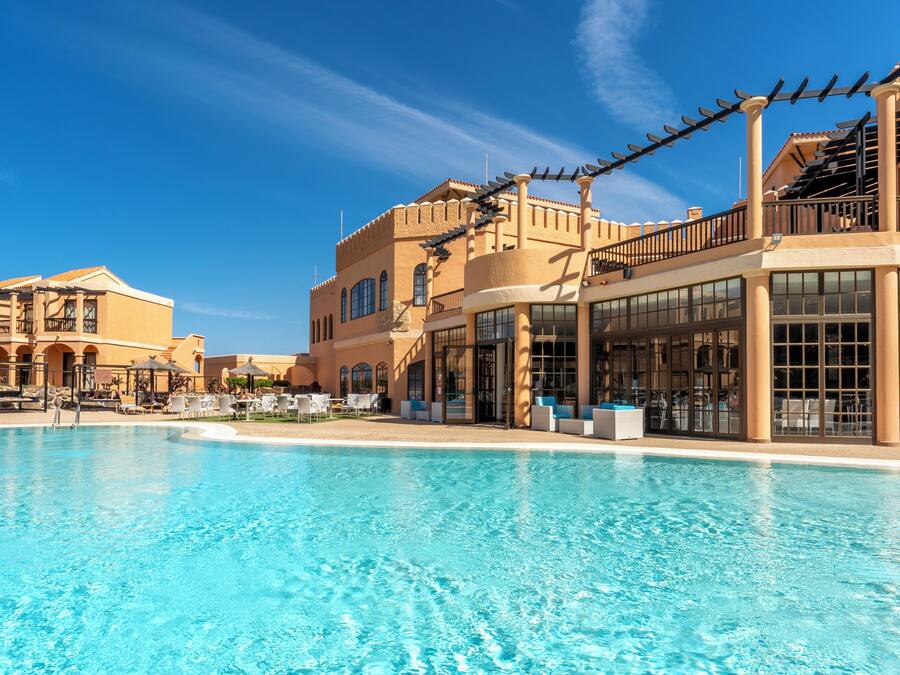 Un resort con una gran piscina, mobiliario exterior y un edificio con varios balcones y ventanas amplias.