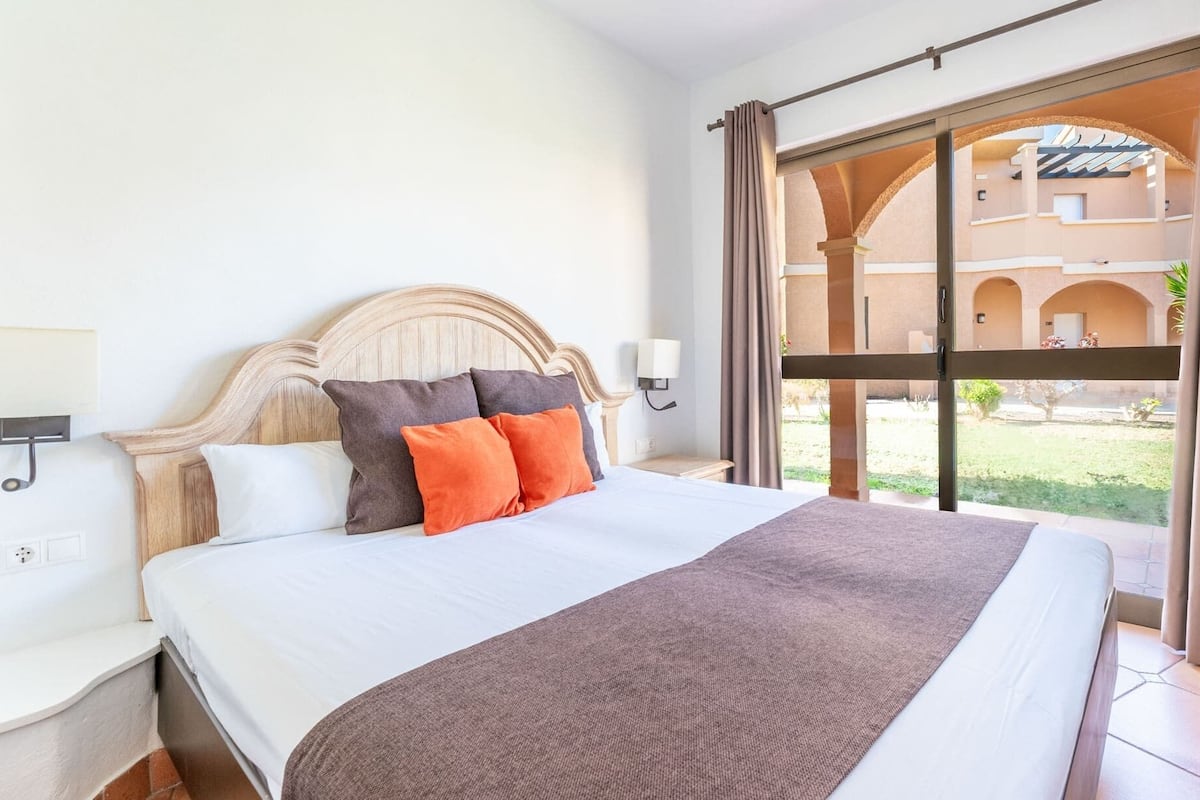 A hotel room with a large bed, a wooden headboard, two bedside lamps, and a view of an outdoor courtyard with arches.