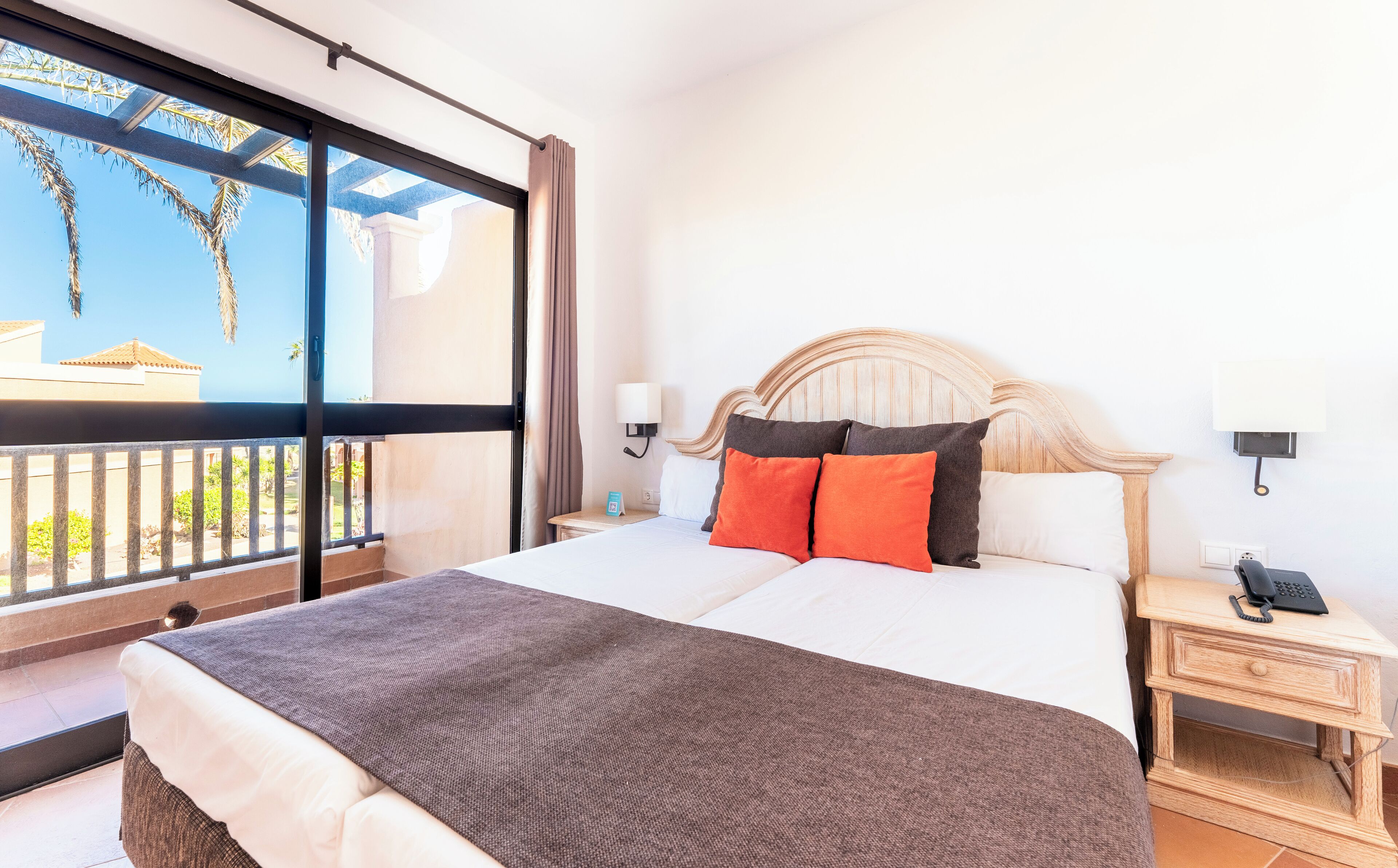 Une chambre d’hôtel avec un grand lit, deux tables de chevet, une petite table et une vue sur les palmiers par une grande fenêtre.