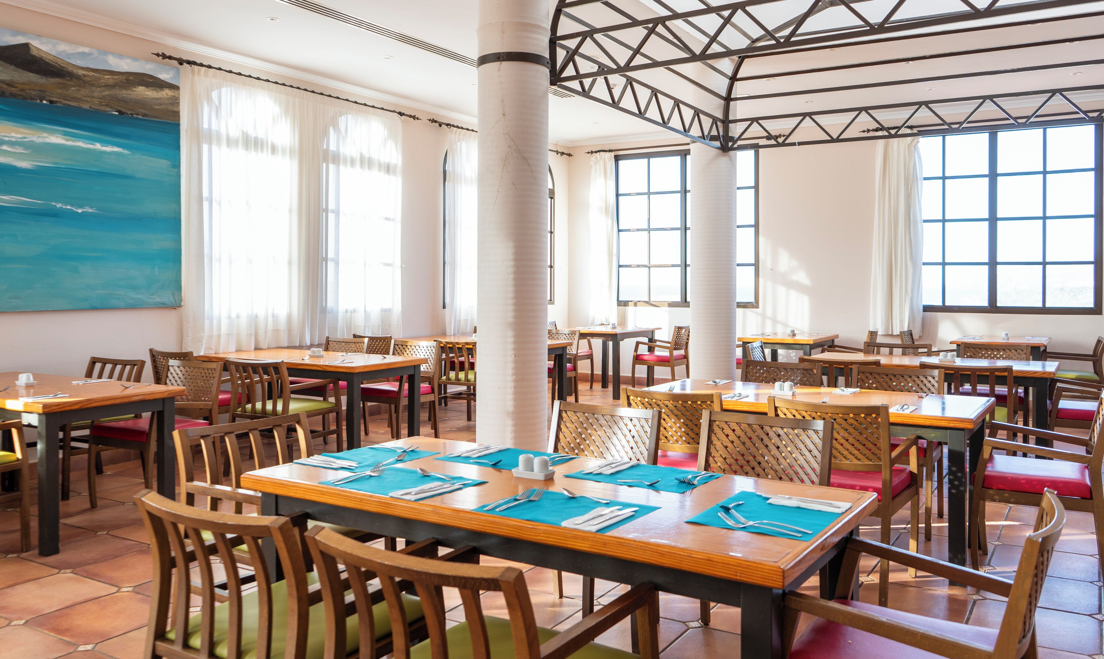 A restaurant with multiple tables and chairs, large windows, and a high ceiling with a metal framework.