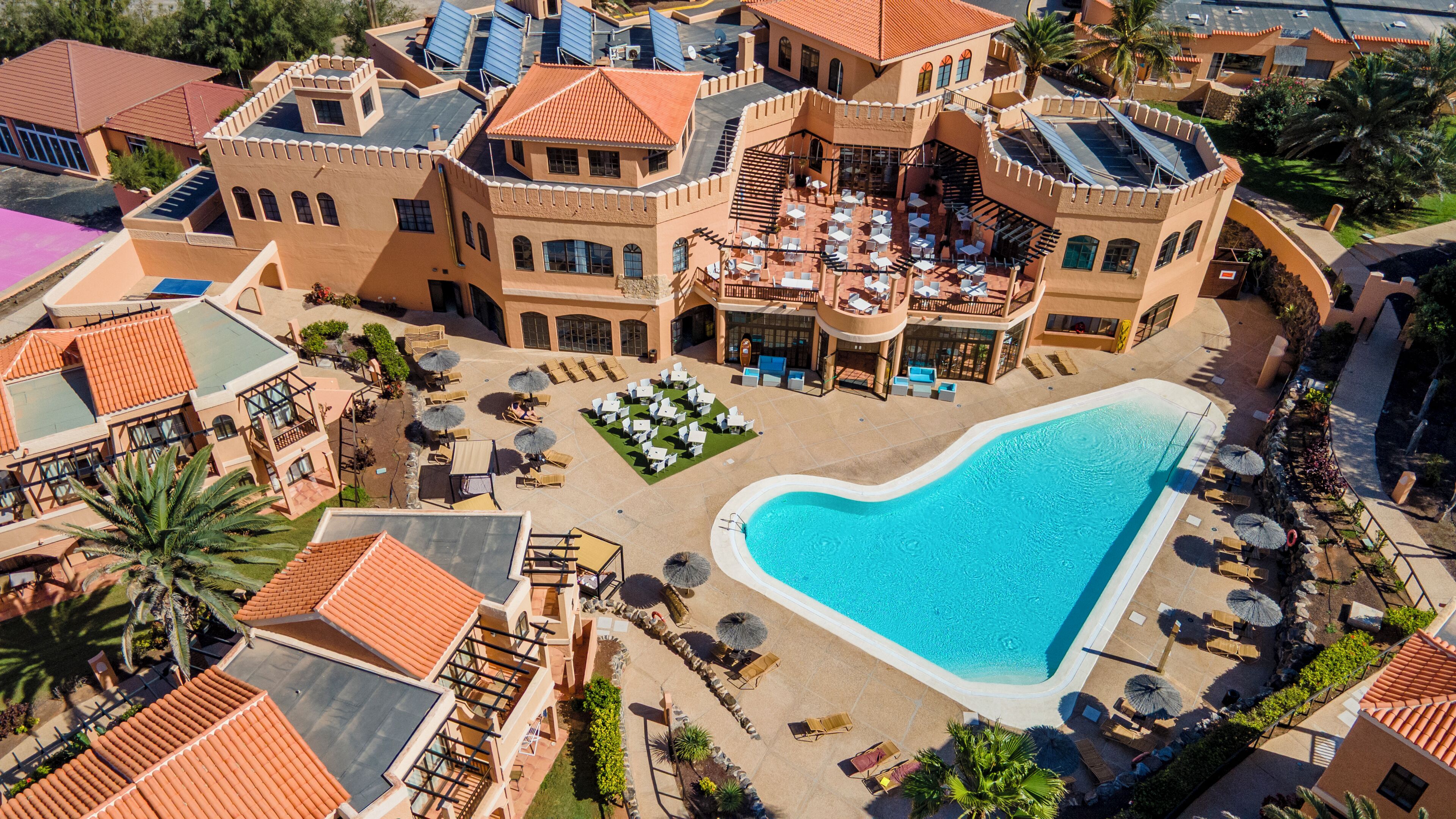 A large resort with a central swimming pool, multiple dining areas, and a variety of outdoor seating arrangements.