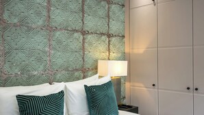 A hotel room with a patterned wallpaper, a bed with white linens and teal pillows, a nightstand with a lamp, and a wardrobe.