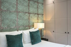 A hotel room with a patterned wallpaper, a bed with white linens and teal pillows, a nightstand with a lamp, and a wardrobe.