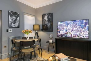 A modern living room with a round dining table, chairs, a vase with flowers, a lamp, a television, and framed artwork on the walls.