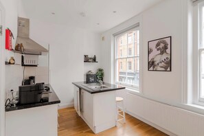 A compact kitchen with white cabinetry, a black countertop, a stainless steel range hood, a window with a view of buildings, and a framed picture on the wall.