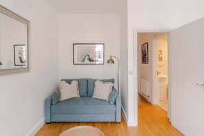 A living room with a sofa, a framed abstract painting, a floor lamp, and a hallway leading to a bathroom.