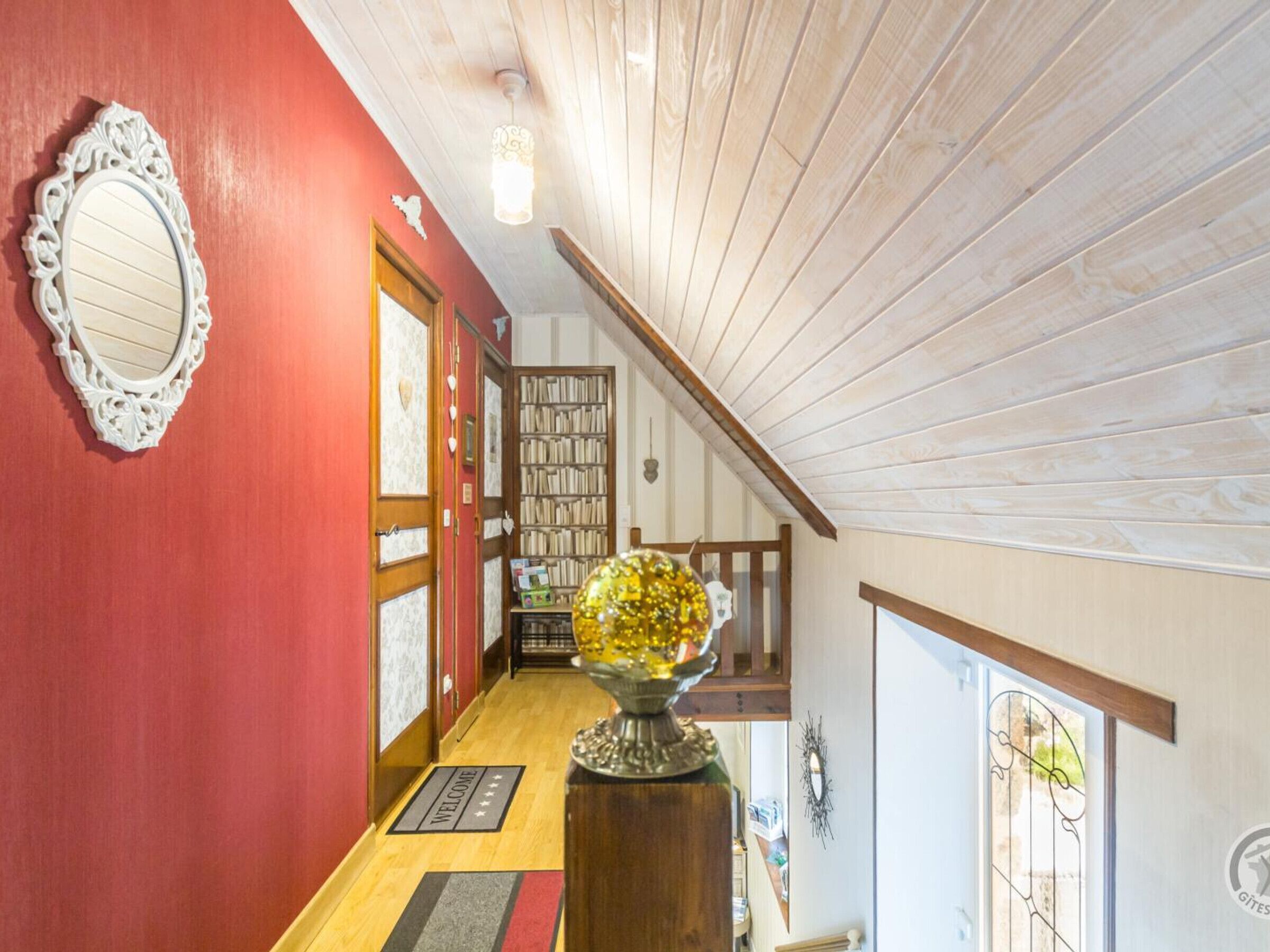 A hallway with a red wall, a white ornate mirror, a wooden door, a bookshelf, and a decorative glass globe on a wooden pedestal.