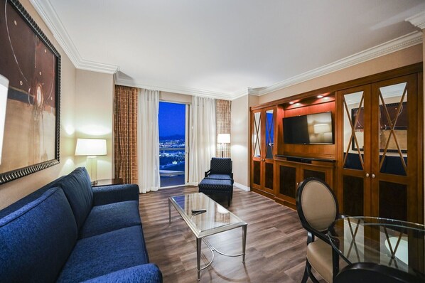 Habitación de hotel con un sofá grande, una mesa de centro de vidrio, un televisor y vistas de la ciudad de noche.
