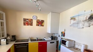 Een keuken met een gootsteen, fornuis en magnetron. Er zijn kastjes, een klok en een raam met zonwering.