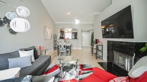 A modern living room with a grey sofa, red and black patterned ottoman, and a glass coffee table. A flat-screen TV is mounted on the wall, and a fireplace is visible in the background. - SVS: Bright 1BD Condo | Gated Community | Pool | Near the Strip (Las Vegas)