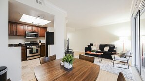 Cozinha moderna com armários de madeira, micro-ondas e fogão. Há uma área de jantar com mesa e cadeiras de madeira, e uma sala de estar com sofá preto e poltrona branca.