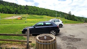 Un’area di parcheggio con due auto ferme vicino a un recinto di legno, circondata da campi verdi e una collina boscosa sullo sfondo.