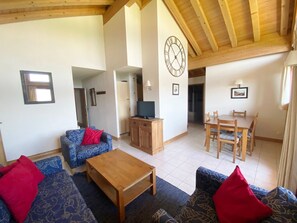 A spacious living area with a wooden ceiling, a dining table with chairs, a sofa set, a TV on a wooden cabinet, and a wall clock.