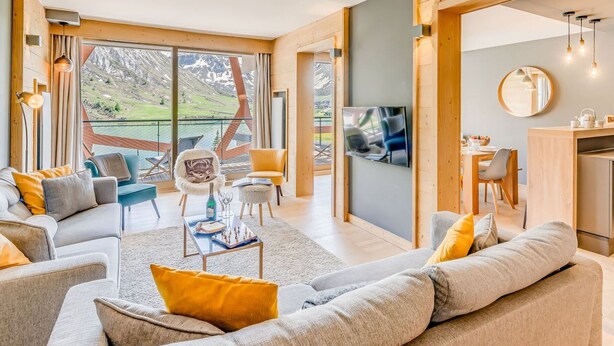 A spacious living room with a large sofa, a dining area, and a kitchen. There is a TV mounted on the wall and a balcony with a view of the mountains.