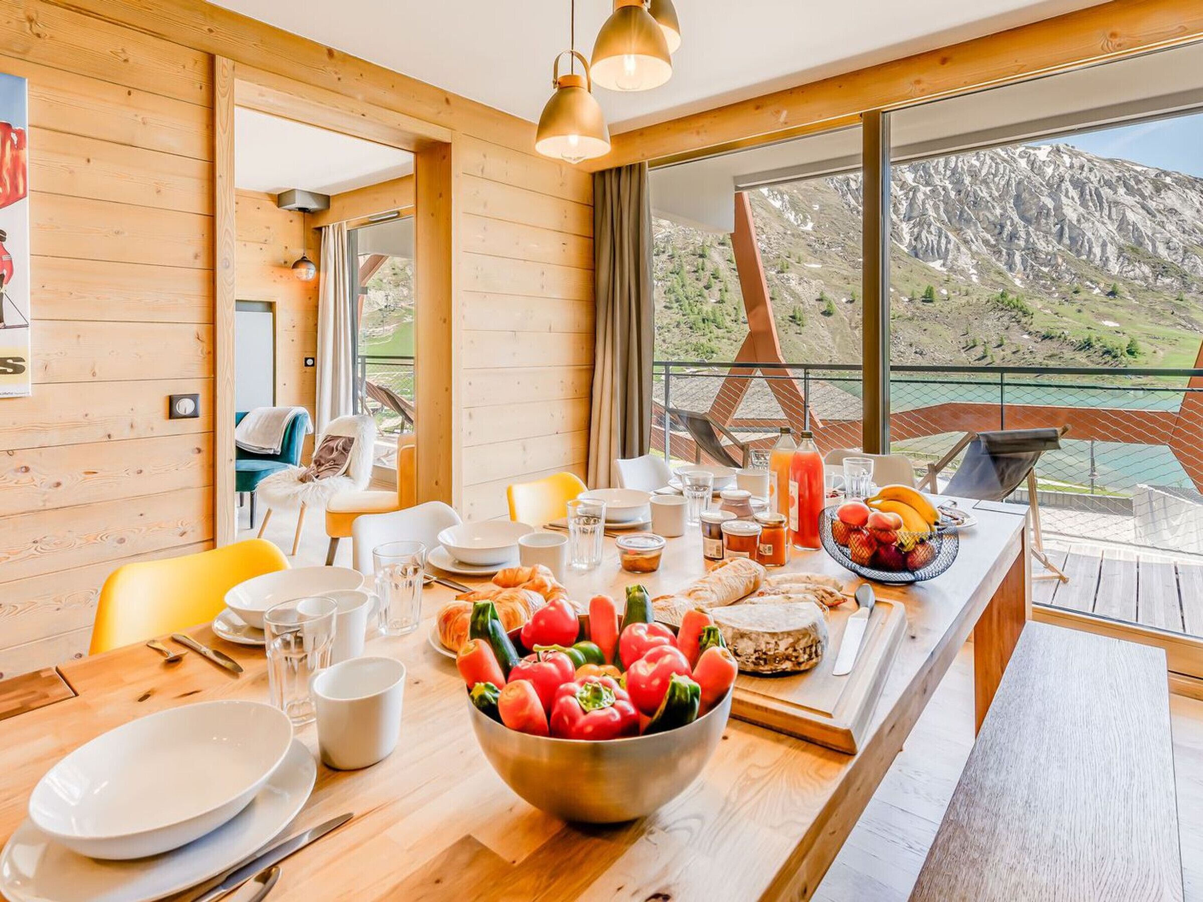 A dining area with a wooden table set for breakfast, featuring a bowl of tomatoes, bread, and a bottle of juice.