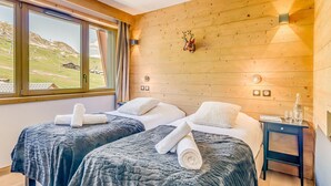 A hotel room with two beds, a wooden wall, a window with a view of a ski slope, and a small bedside table with a bottle and glasses.
