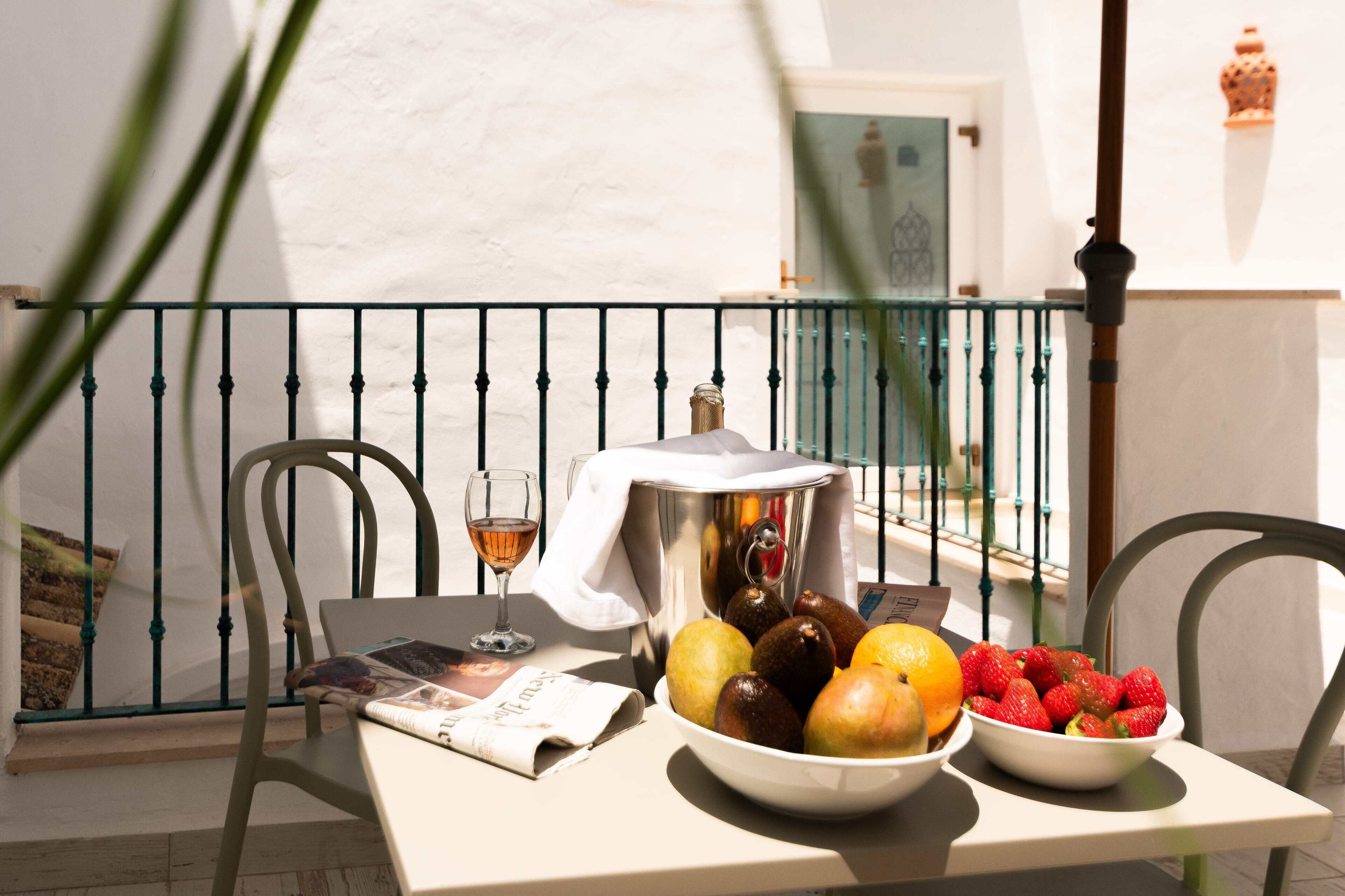 A table with a newspaper, a glass of wine, a bowl of fruit, and a metal container on a balcony with a railing.
